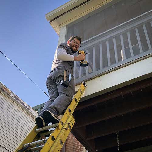 A man holding a drill is perched atop a ladder. He is smiling.